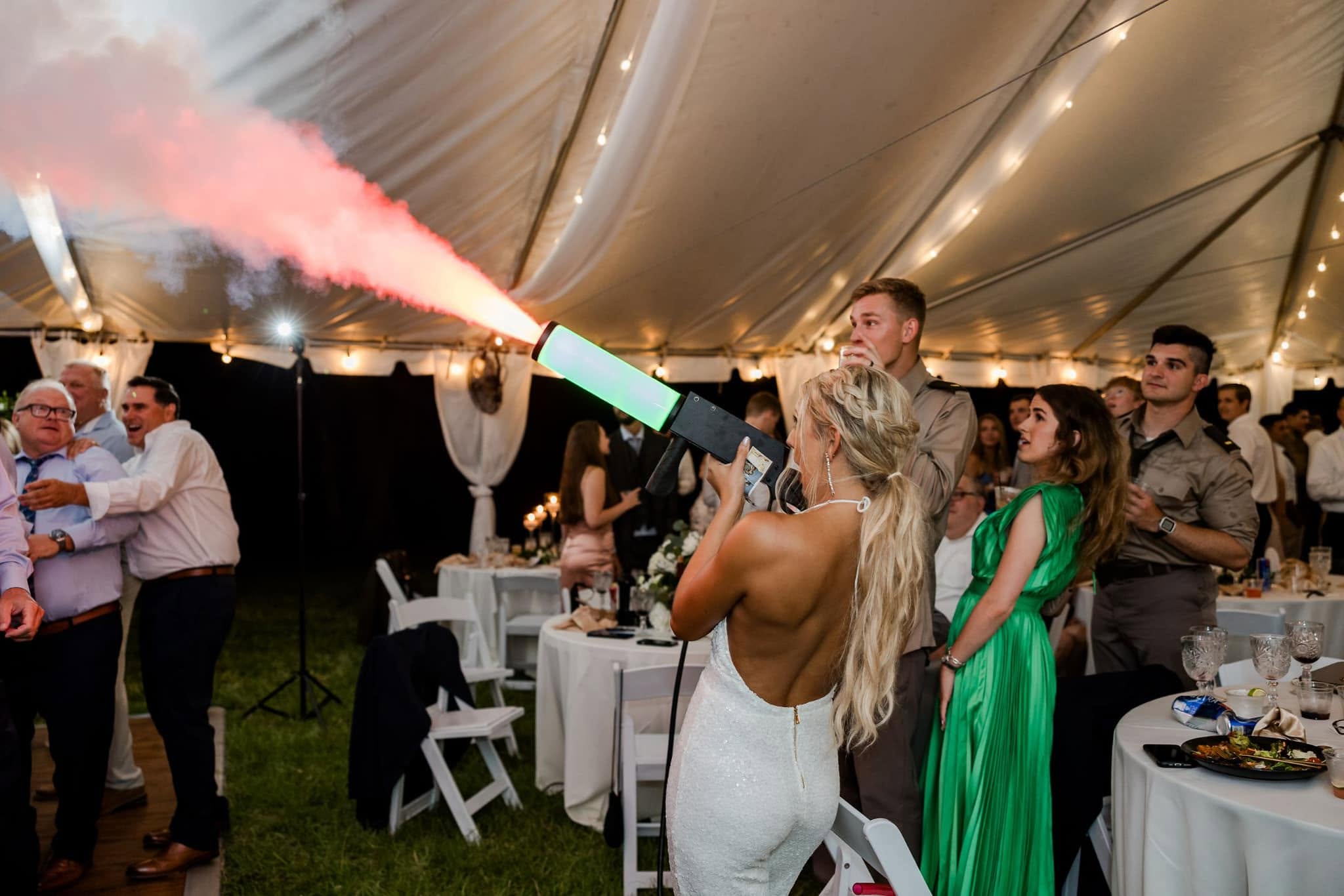 Bride in a white dress stands with her back to the camera as a pink smoke flare shoots into the air at a wedding reception under a string-lit tent.