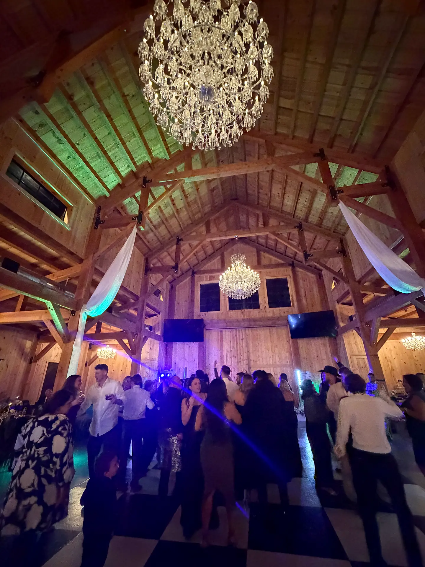 Guests dancing on a checkered dance floor beneath crystal chandeliers and colored lights in a barn venue in Alva