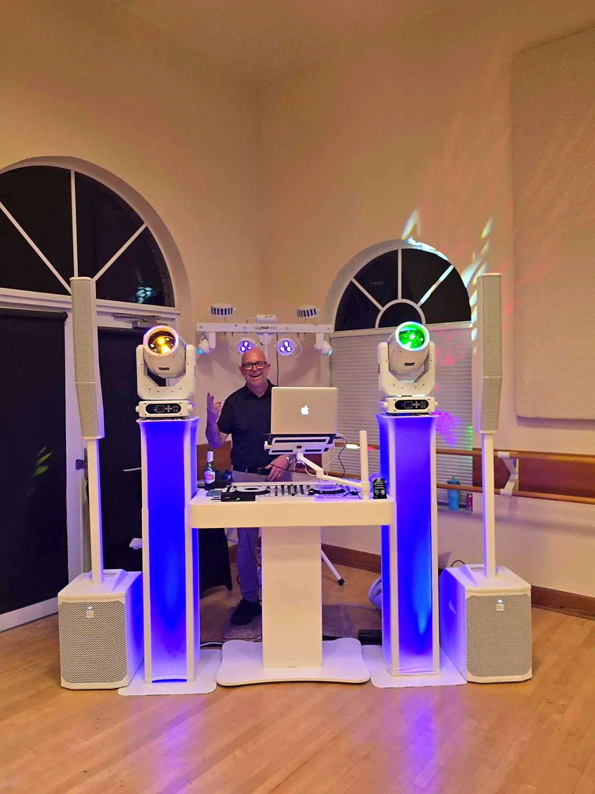 DJ Bigs behind a white DJ booth with blue uplit totems, moving-head lights and speakers in a ballroom with arched windows