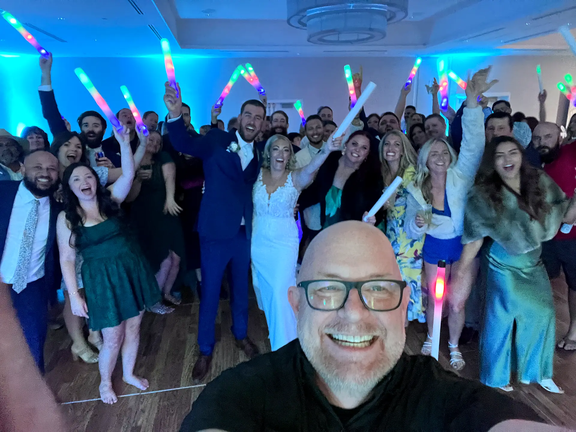 DJ Bigs takes a selfie with the bride, groom and guests waving LED foam glow sticks at a Fort Myers Beach wedding reception
