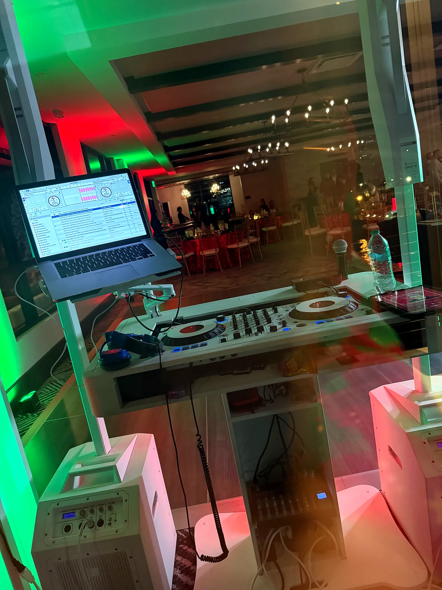 View from behind the decks of a DJ controller and laptop overlooking a red and green lit reception in Fort Myers