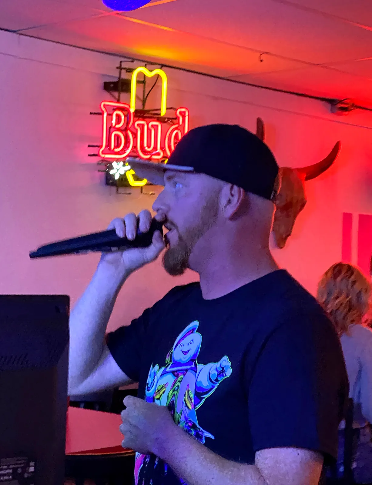 A singer in a black cap on the mic under a red neon beer sign at a Southwest Florida karaoke night